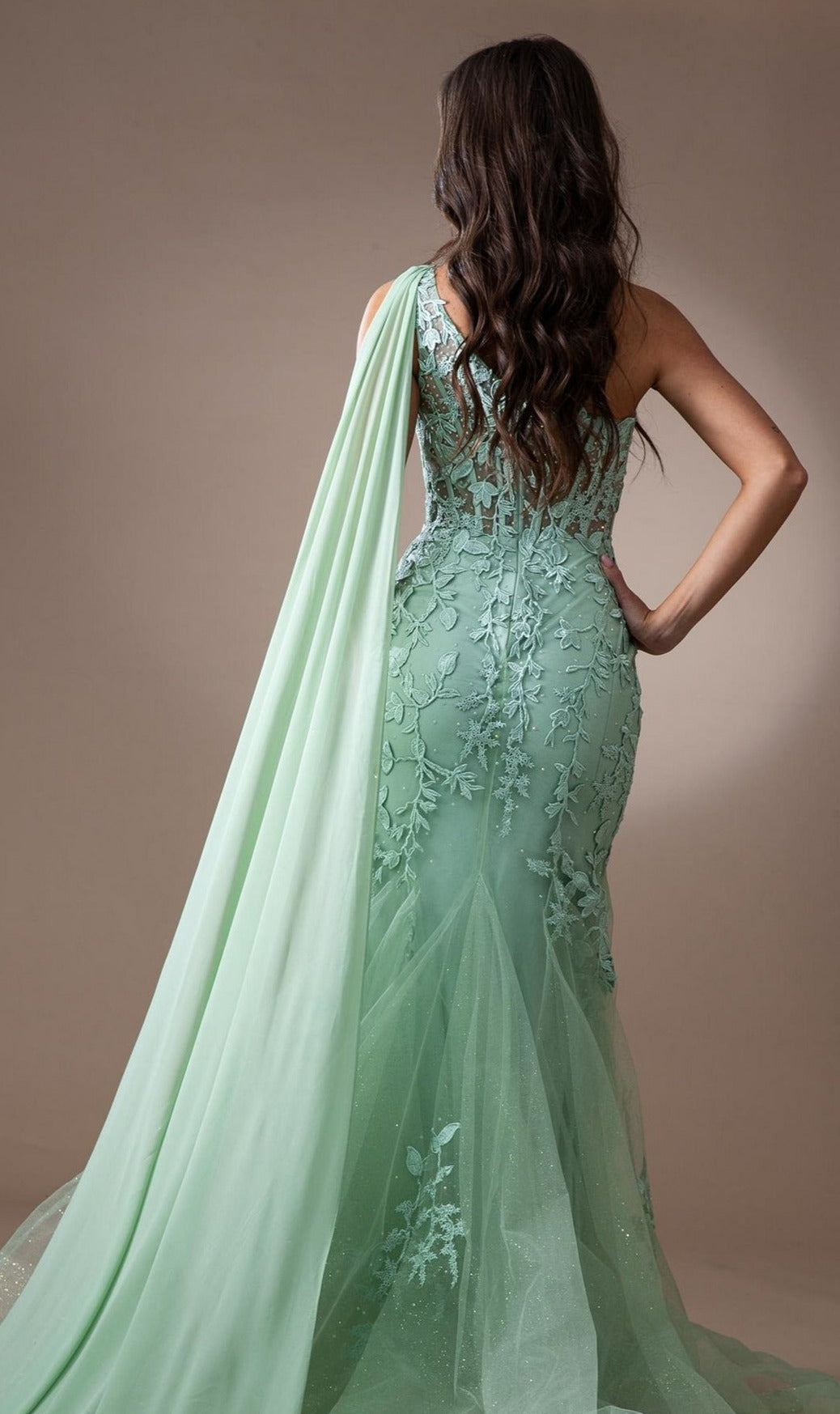 A woman stands facing away, wearing the One-Shoulder Long Lace Mermaid Prom Dress 7048 in mint green, featuring floral lace, a fitted floor-length silhouette, and a long sash draped over one shoulder.